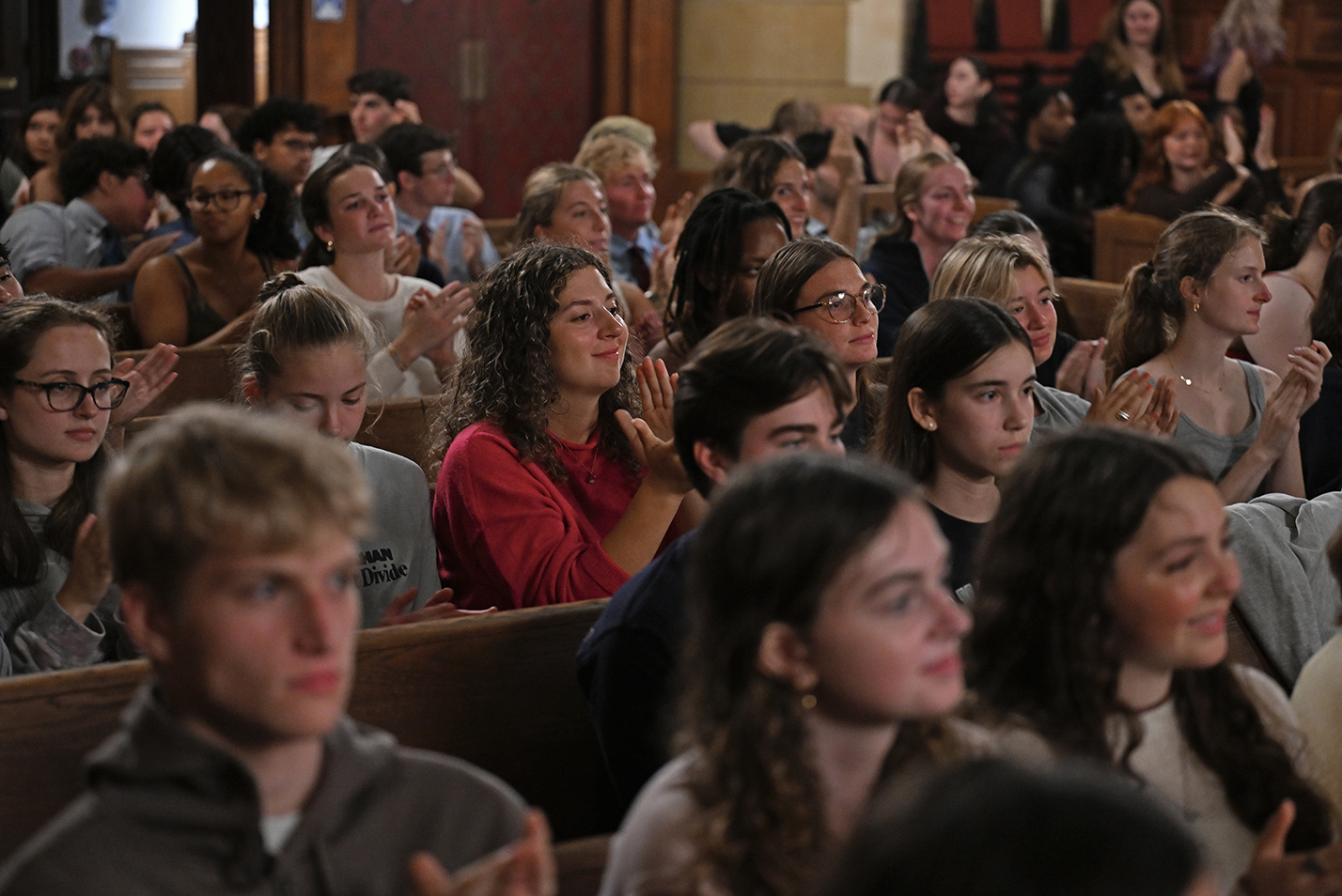 An audience of college students seated in the pews of a chapel applaud an a cappella performance.