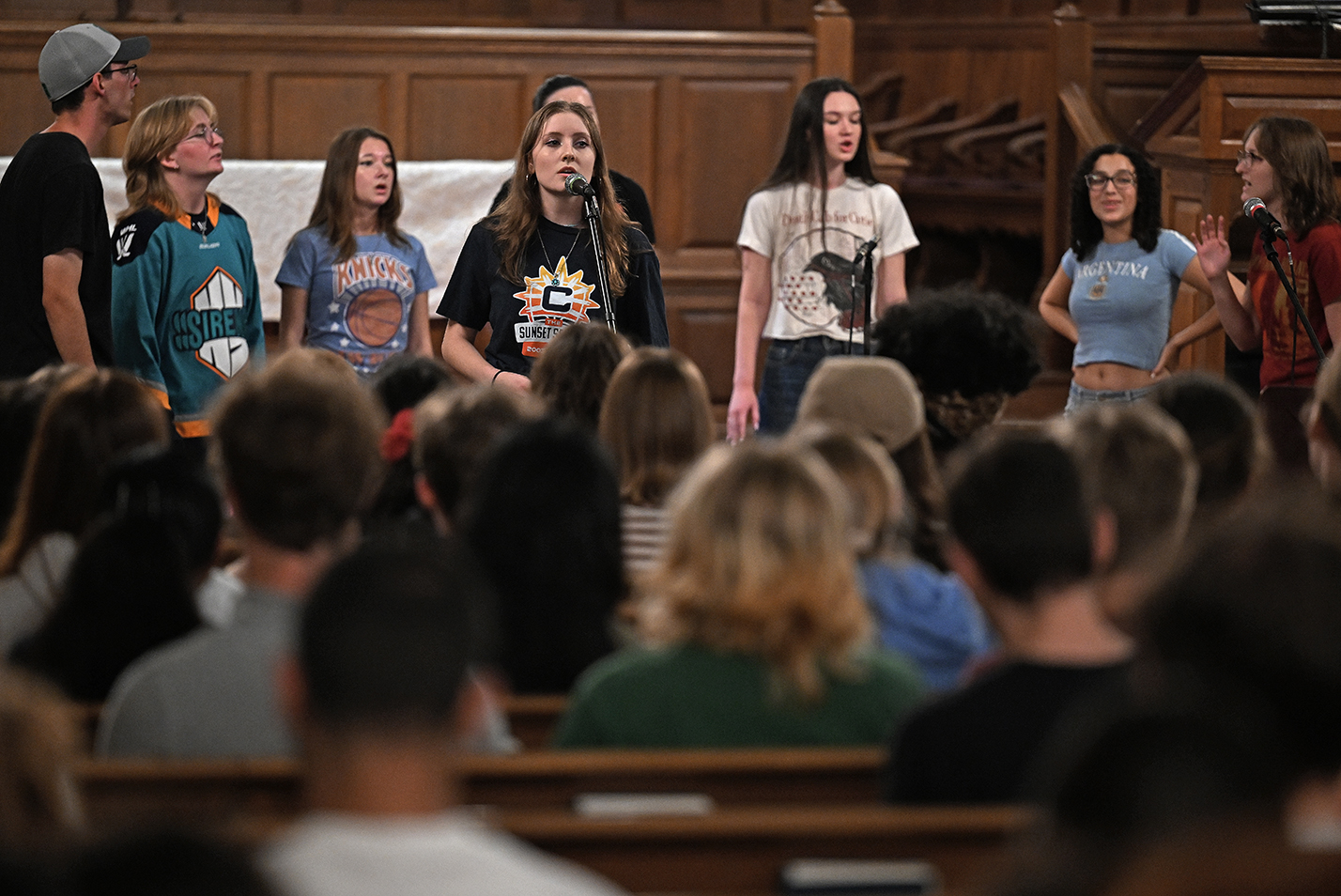 A college a cappella group wearing sports team shirts performs in front of an audience seated in the pews of a chapel.