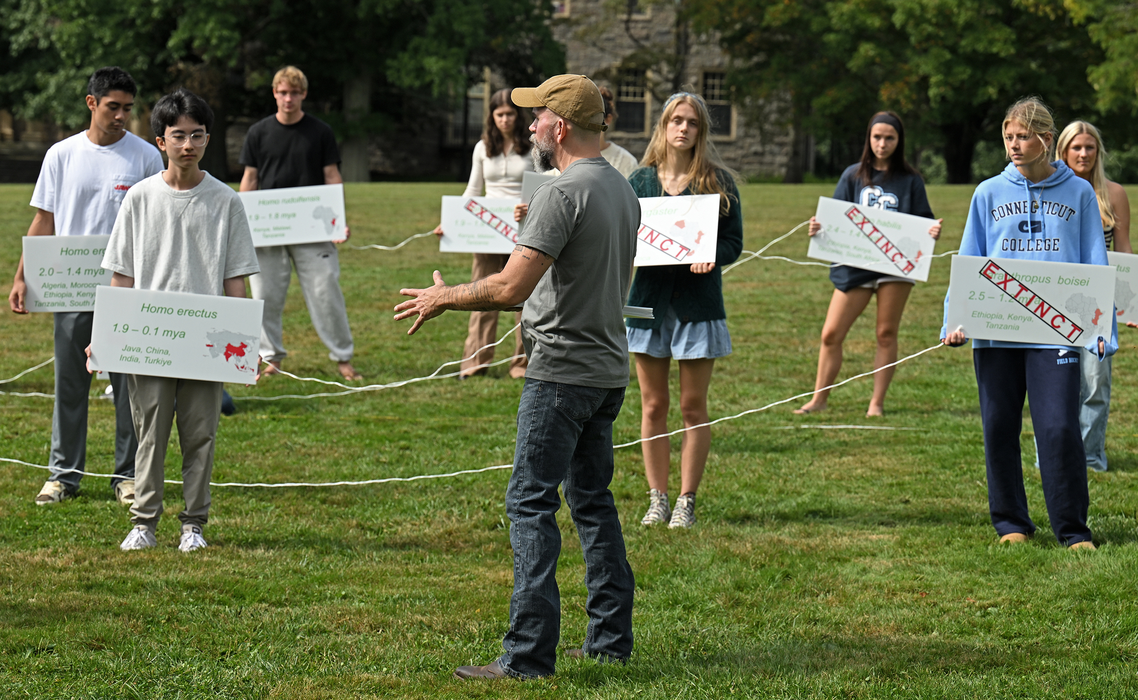 A college professor in blue jeans and a khaki ball cap speaks to students standing out on a green holding signs printed with names and information on pre-homo-sapien ancestors.