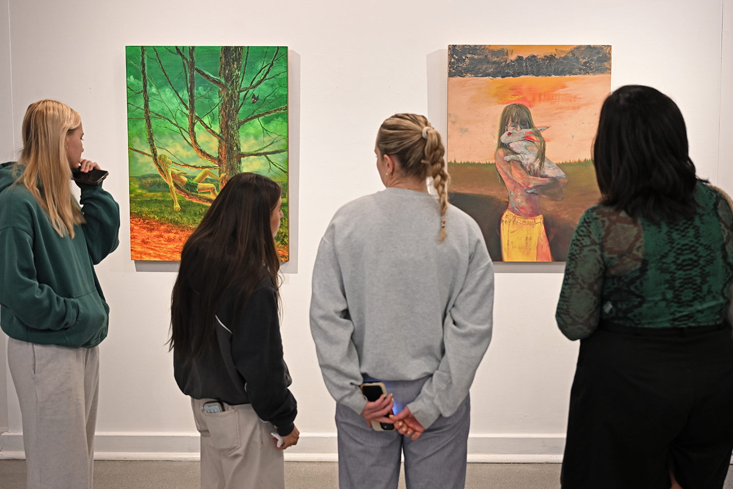 Four visitors to an art gallery view three oil paintings on the wall.