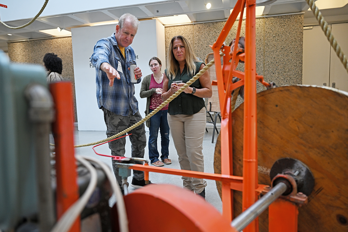 An artist in blue plaid shirt points at a piece of industrial art featuring orange wheels and pipes, and rope, as he explains the piece to two women looking at it in an art gallery.
