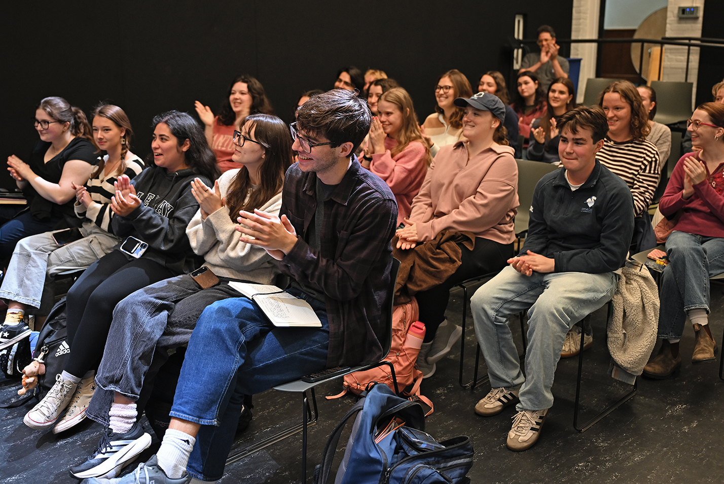 An audience of college students applaud guest speakers not visible in the frame during a question and answer session with broadway performers in a small theater rehearsal space.
