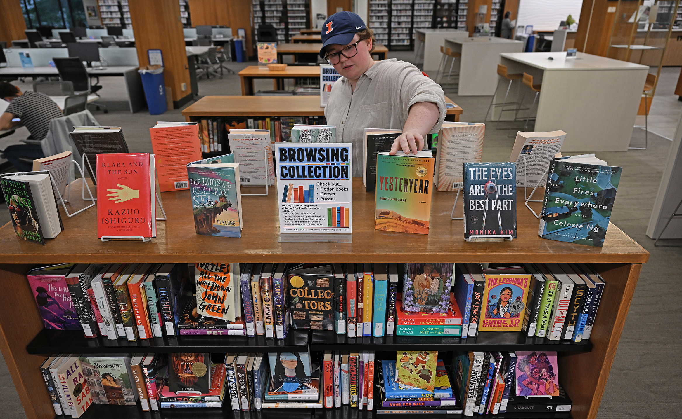 A college librarian wearing a ball cap with a large orange I carefully arranges hardcover books atop a shelving unit.