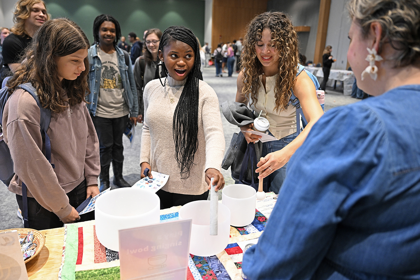 Three female college students smile at the chaplain as they experience ringing a glass singing bowl on a table in a ballroom.