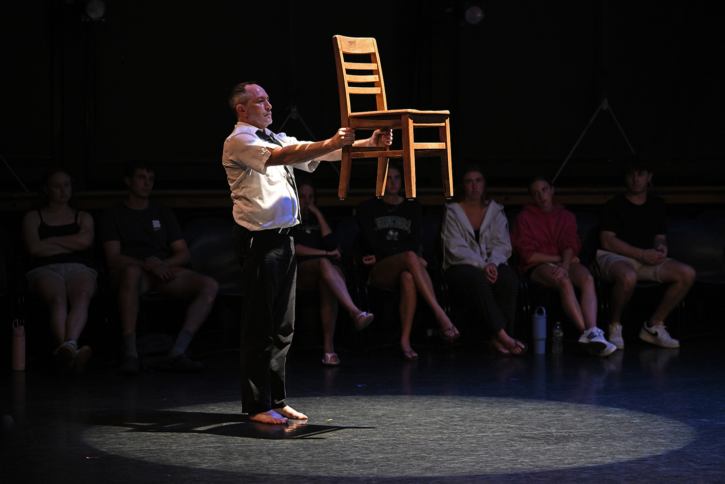 A dance performer in shirt and tie and pants with bare feet holds a chair out at arms length while illuminated by a spotlight and in front of a single row of audience.