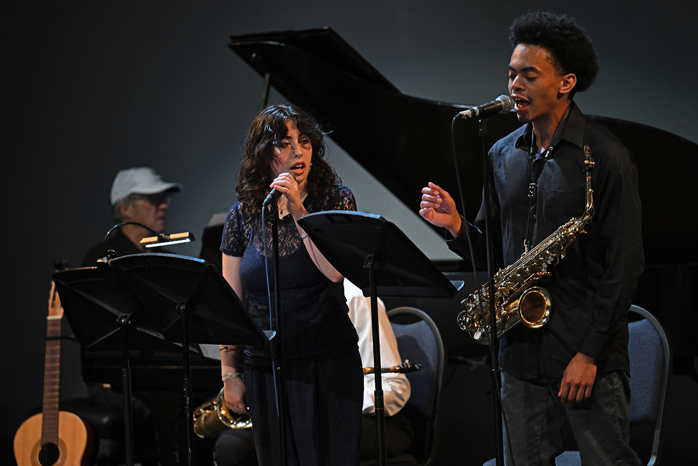 Two singers, one a young woman in a dark outfit with long curly hair and the other a black man with a saxophone hanging from a neck strap sing while accompanied by a pianist in the background.