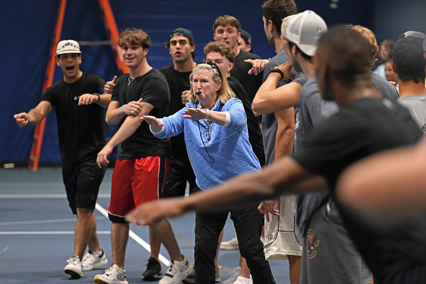 A college athletic director in blue sweatshirt blows a whistle and gestures to make a call surrounded by shouting and gesturing male student athletes on the sidelines of a dodgeball tournament.