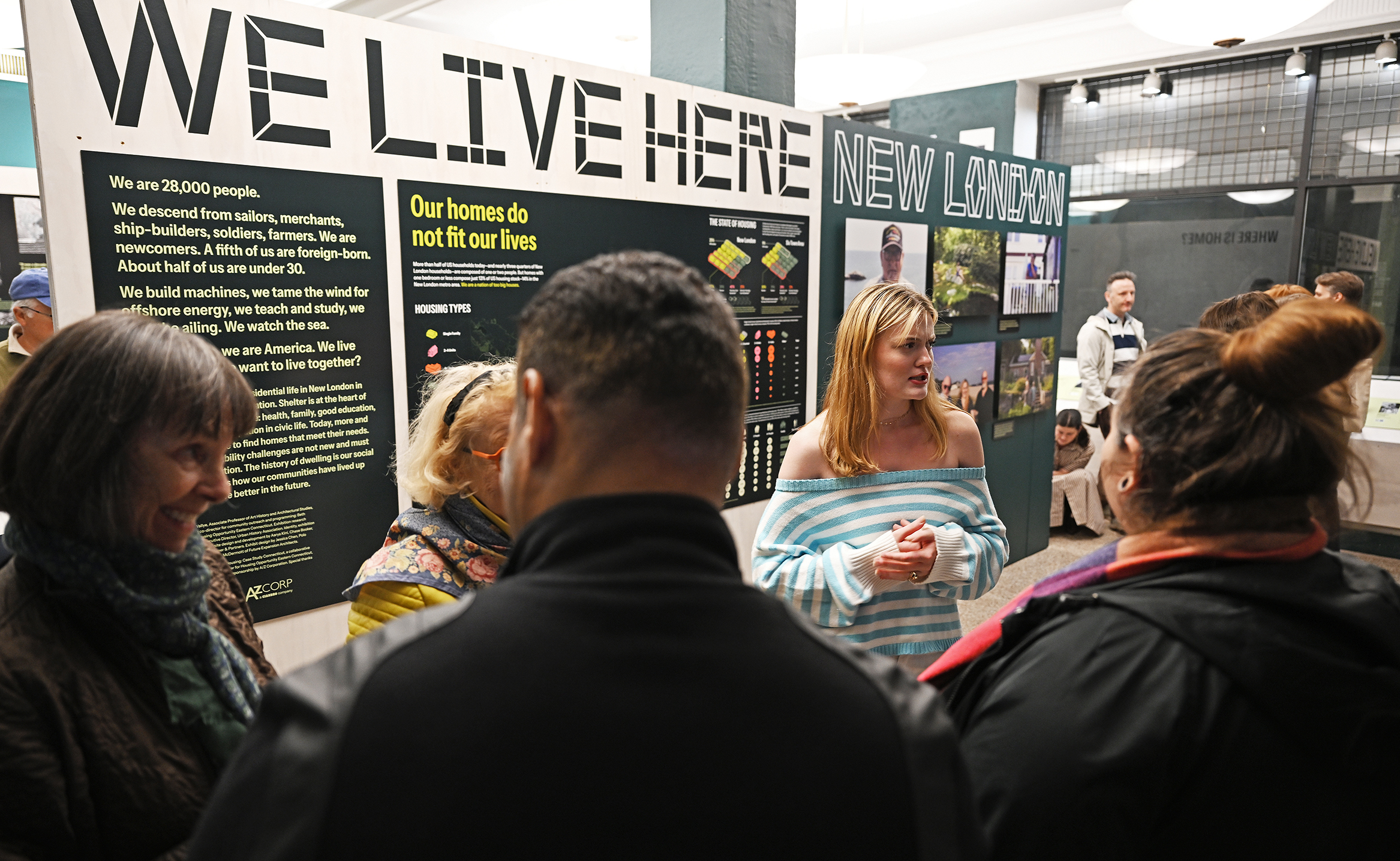 A blond-haired female college student in an off-the-shoulder light blue striped shirt speaks to visitors to a gallery with a sign reading 