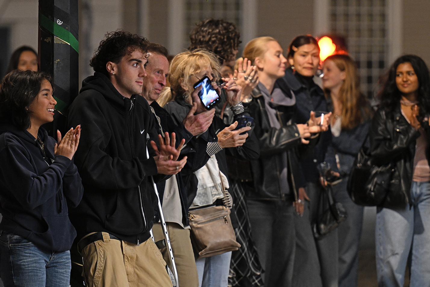 A crowd standing on a sidewalk at night applauds a band playing out of frame.