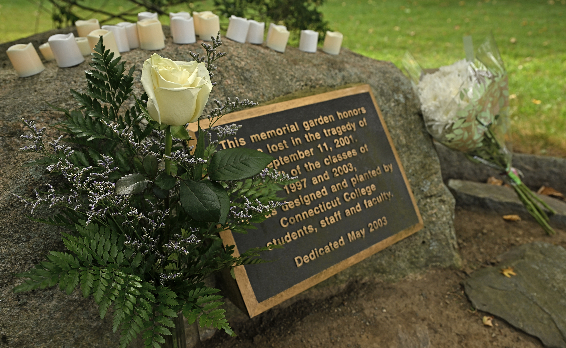 A white rose and a white carnation and a row of electronic candles adorn a 9/11 memorial plaque mounted on a stone in an outdoor memorial garden on a college campus.