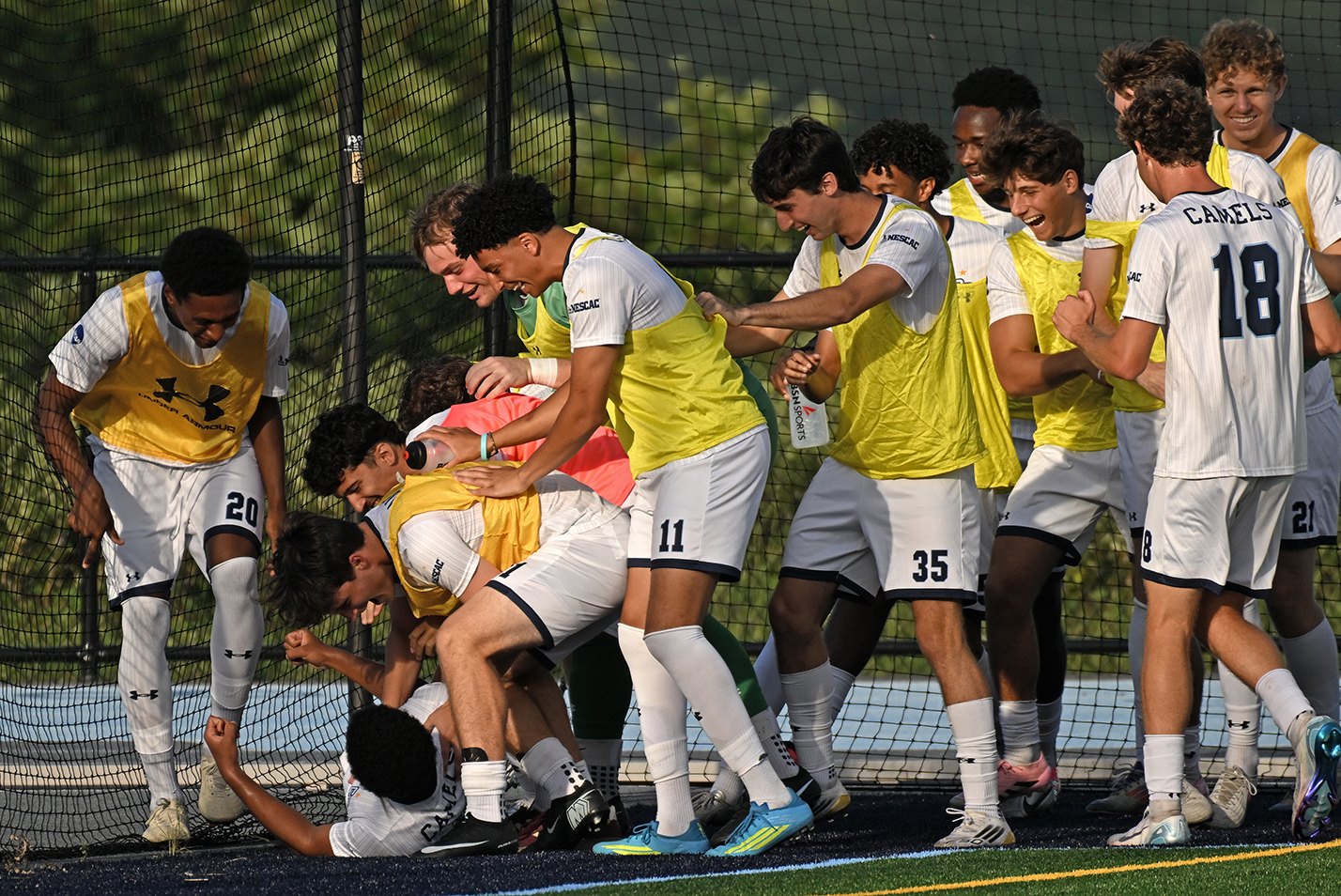 college men's soccer players, many in yellow sideline pennies, mob a teammate on the ground after a goal.