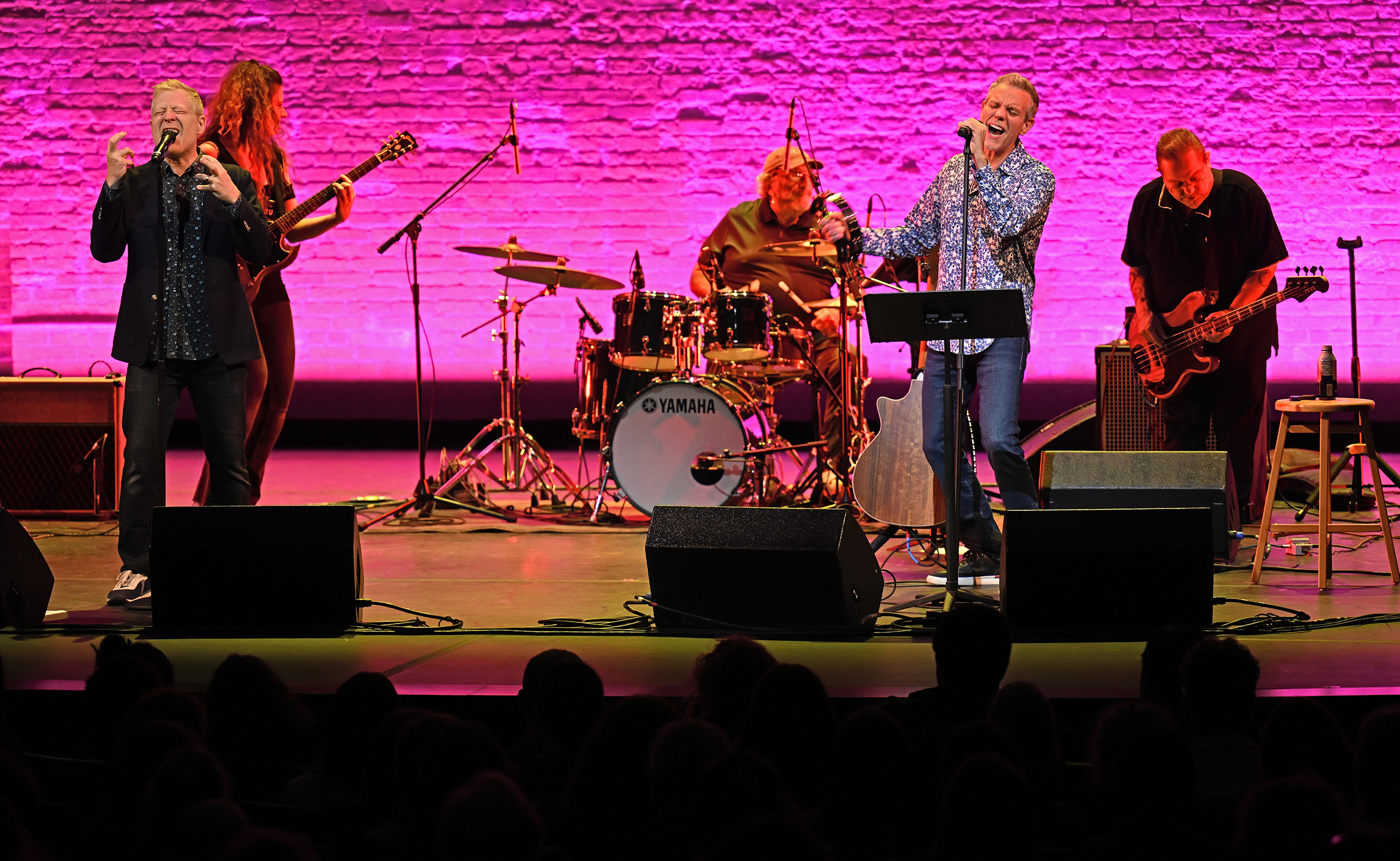 A rock group, fronted by two male vocalists, performs on stage against a brick wall backdrop lit bright pink.