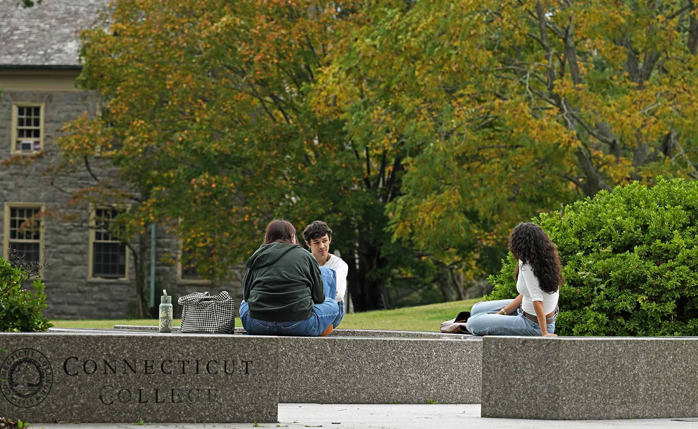 Three college students sit on granite benches on a campus green with changing fall colors visible in the distance.