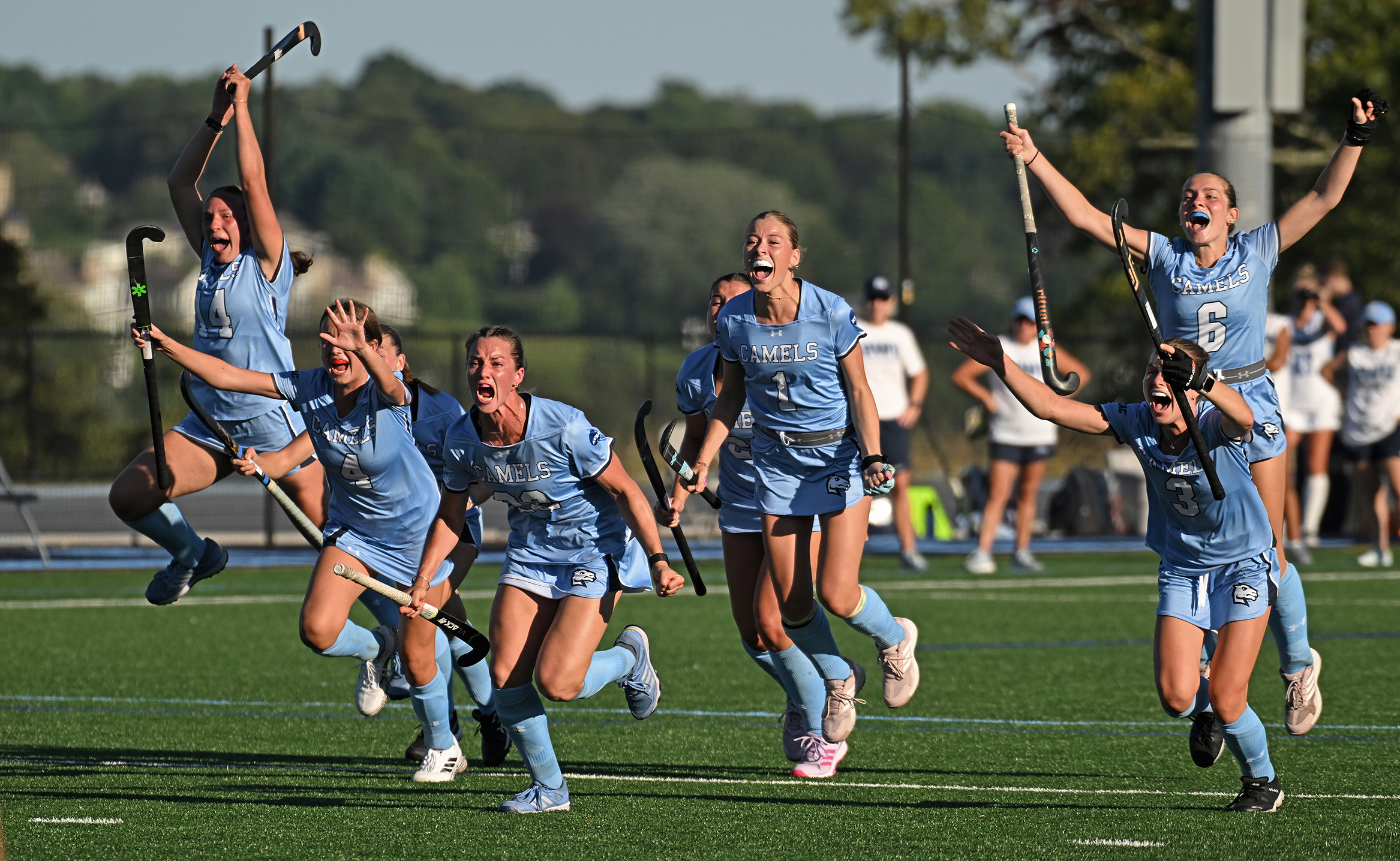 Field hockey players in light blue uniforms cheer and jump in the air as they run across a turf field to celebrate a goal.