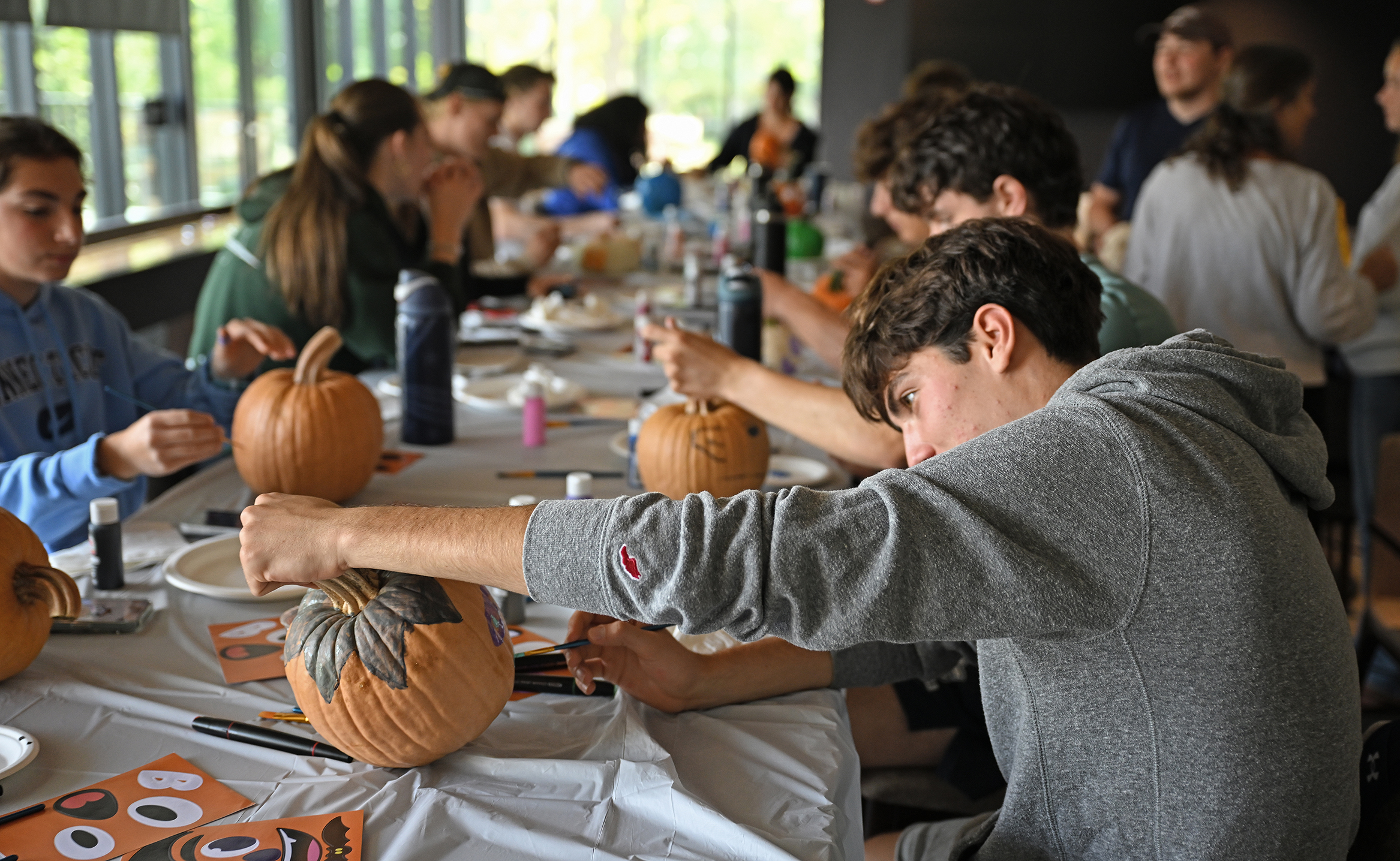 A college student in a grey hooded sweatshirt examines a small pumpkin he is painting on a dropcloth covered table lined with other students also decorating pumpkins.