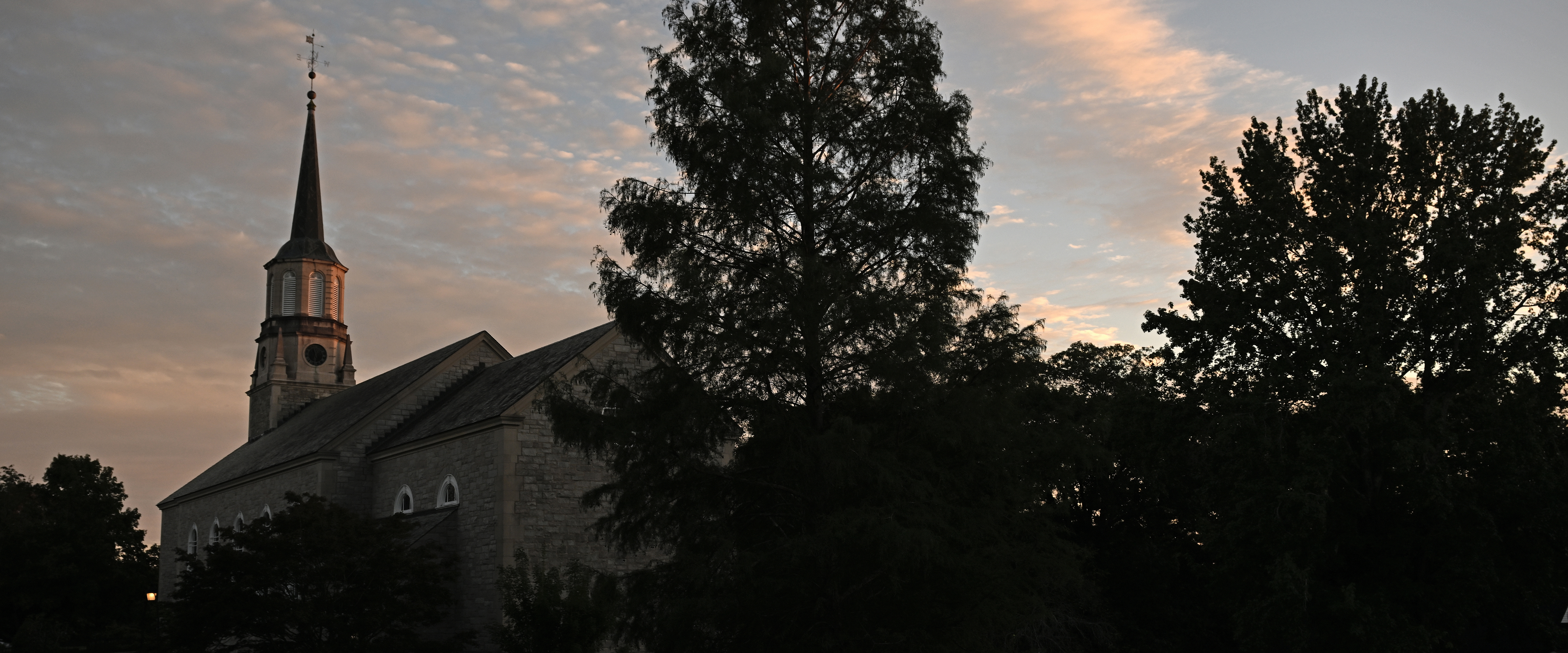 A church steeple and a tree are silhouetted against a sky of clouds glowing yellow/orange from the setting sun.