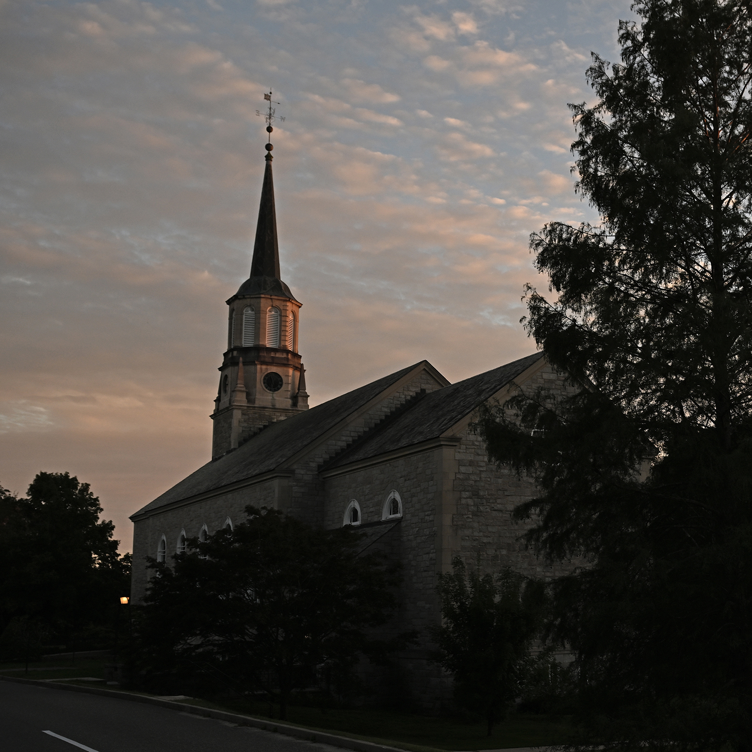A church steeple and a tree are silhouetted against a sky of clouds glowing yellow/orange from the setting sun.