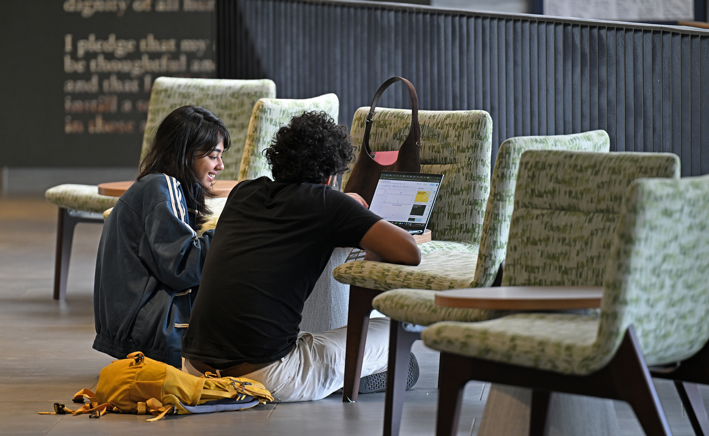 Two college students sit on the floor in a student union space with a laptop on the chair in front of them.