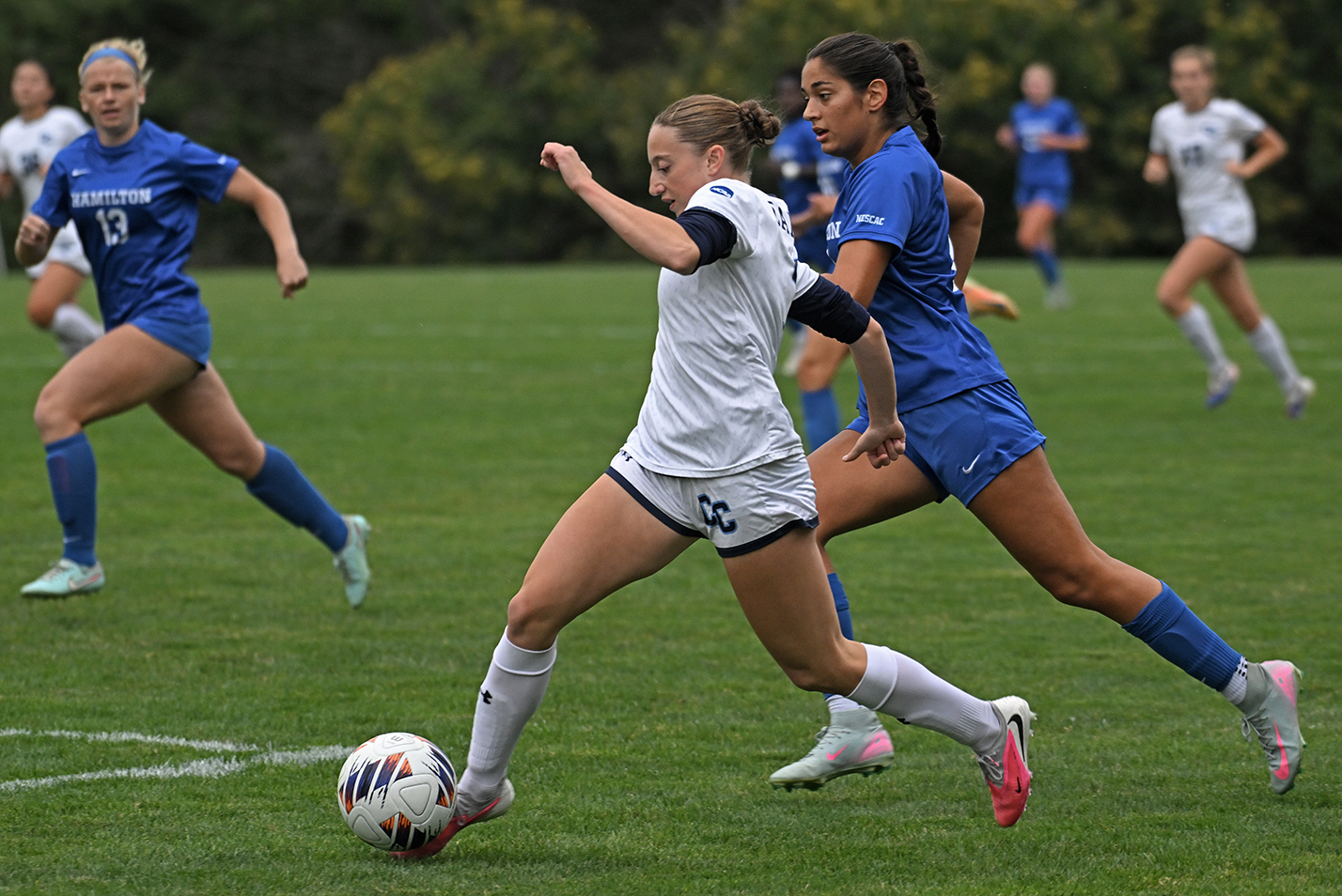 A women's college soccer player in white uniform dribbles the ball with her toe while being pursued by an opponent in a blue uniform.