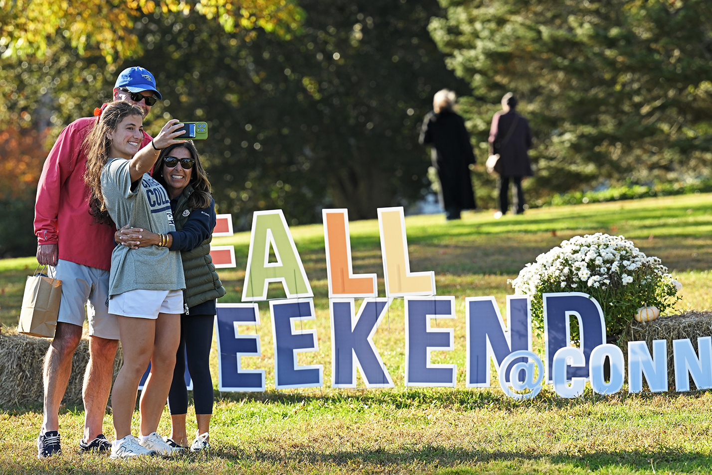 A college student and her parents pose for a selfie-style photo in front of a lawn display readings 
