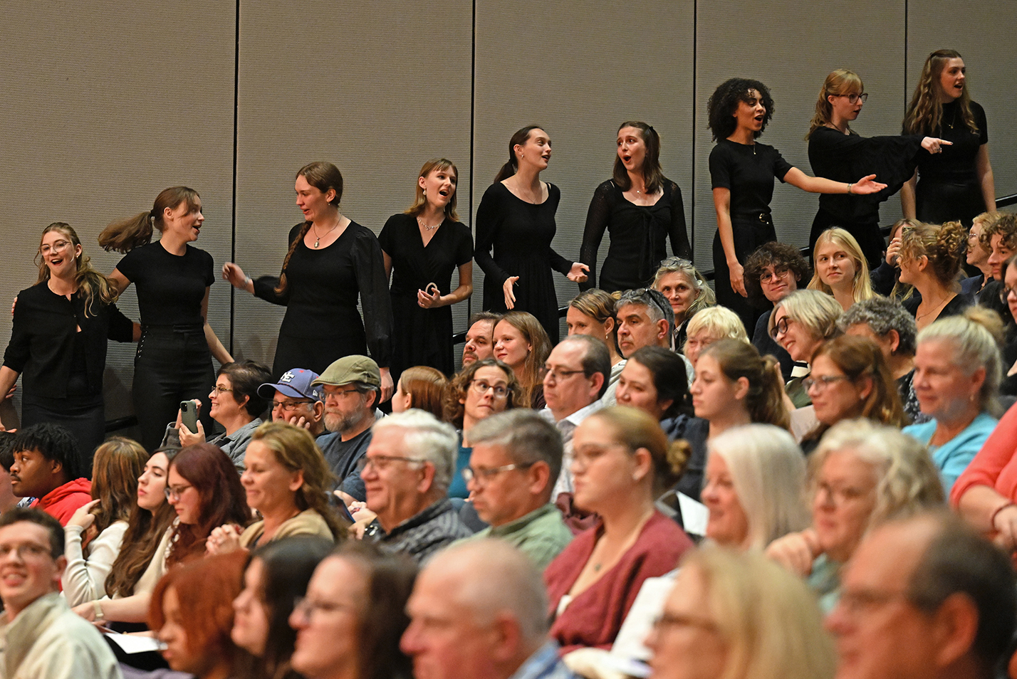 Female members of a college choral singing group dressed in black sing while standing in the aisle by a full-house crowd seated in a steep performance hall auditorium.