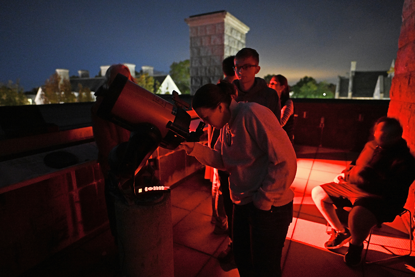 People peer through a reflecting telescope on a rooftop terrace illuminated by red lights under a night sky.