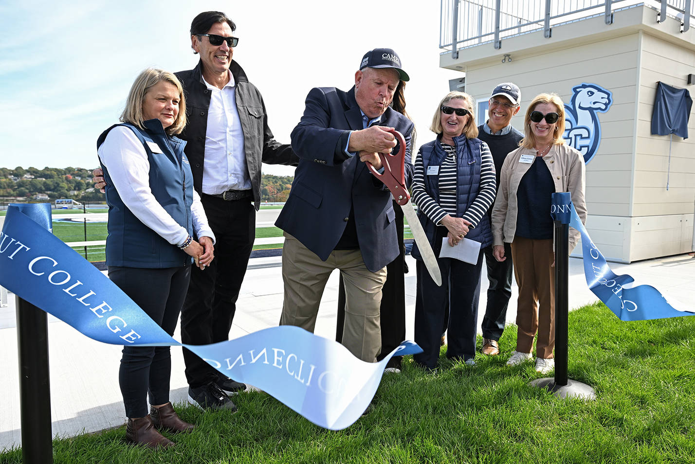 A group of college officials, with one in the middle holding giant ceremonial scissors, cut the ribbon for a new athletic facility.
