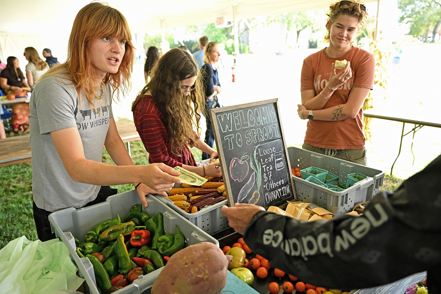 A college student with long red hair hands change back to a customer at a farm stand set up under a festival tent.