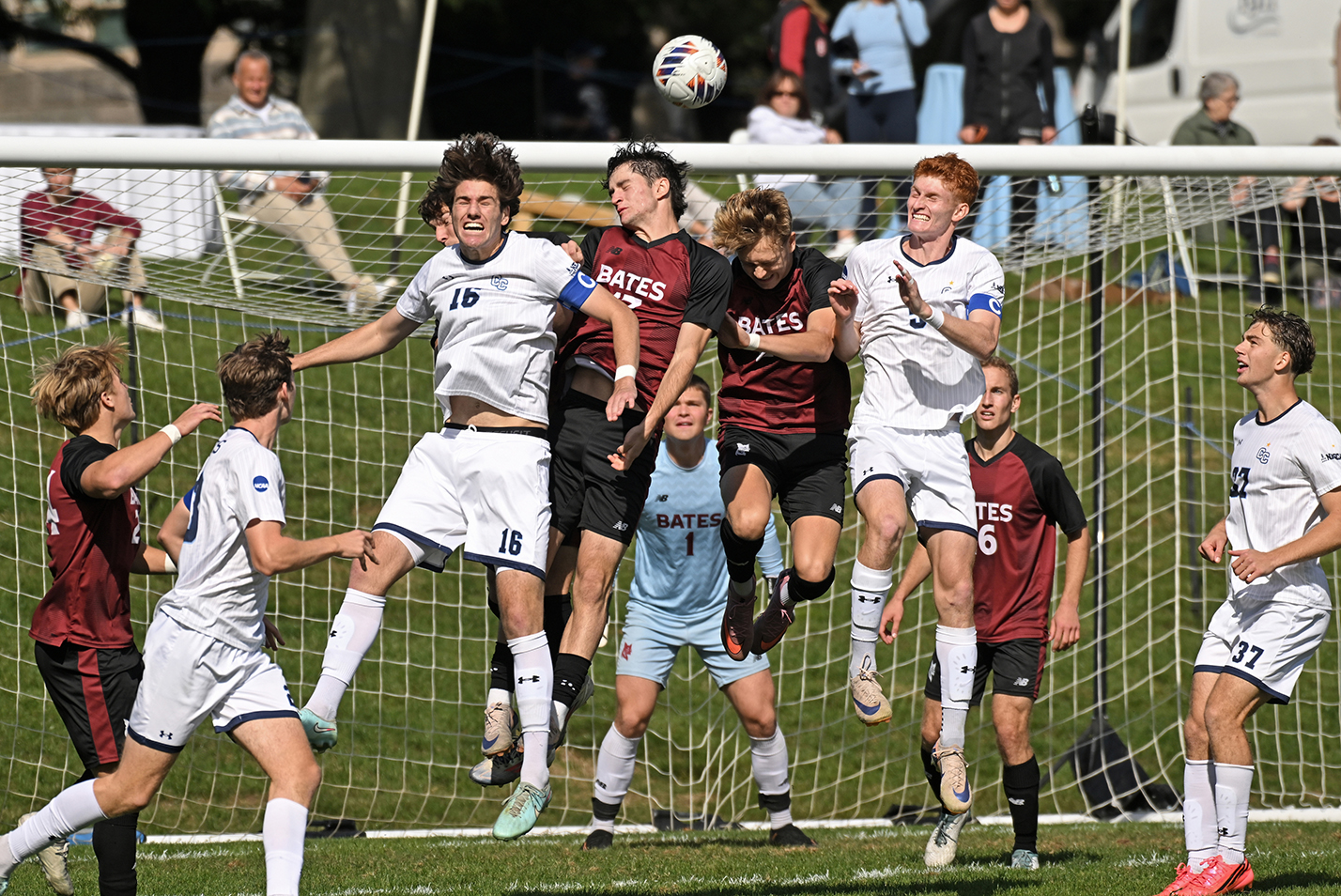 Male college soccer players jump in the air to attempt to head the ball in front of the mouth of the goal.
