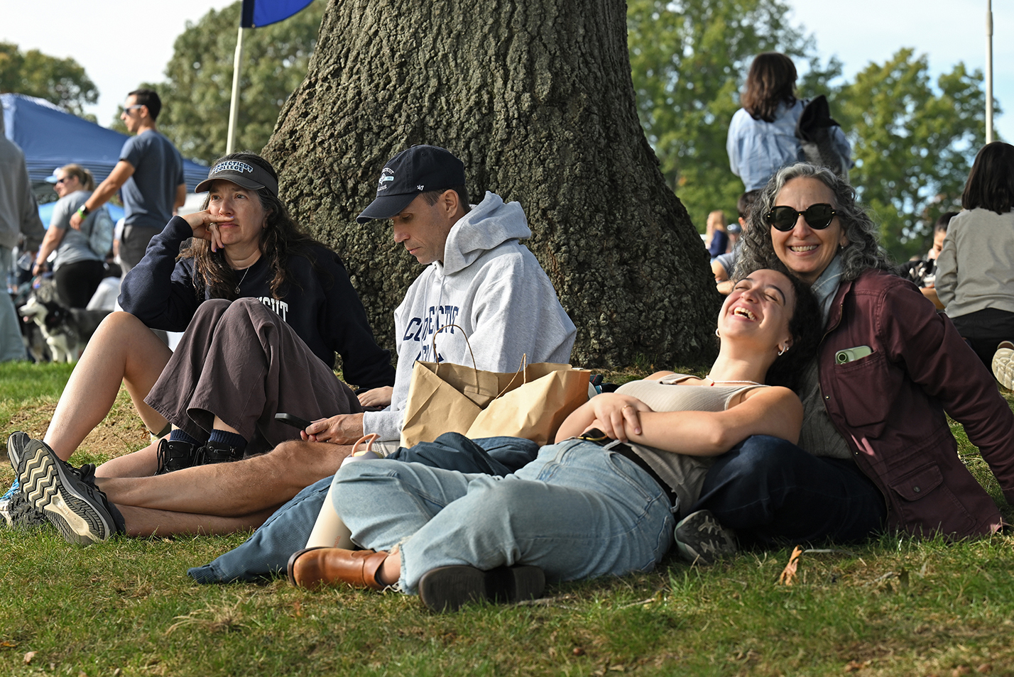 A young woman reclining on the lap of her parent smiles while relaxing on the lawn under an oak tree surrounded by a crowd watching a soccer game.