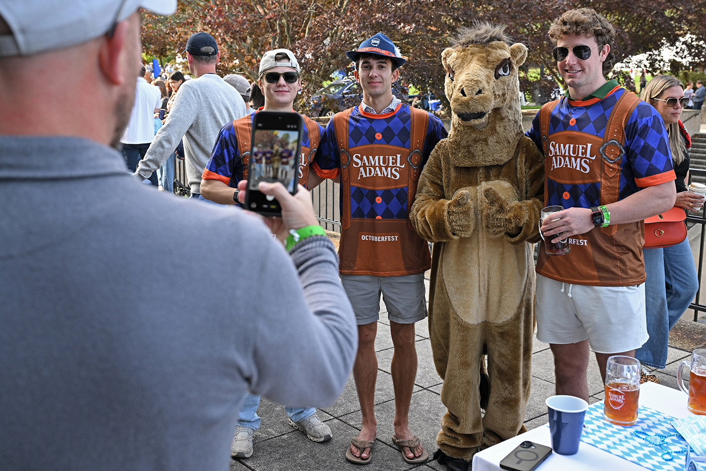 Three young men in faux lederhosen costumes labeled with a beer brand name give thumbs-up signs while posing for a photo with a college mascot in a camel costume.