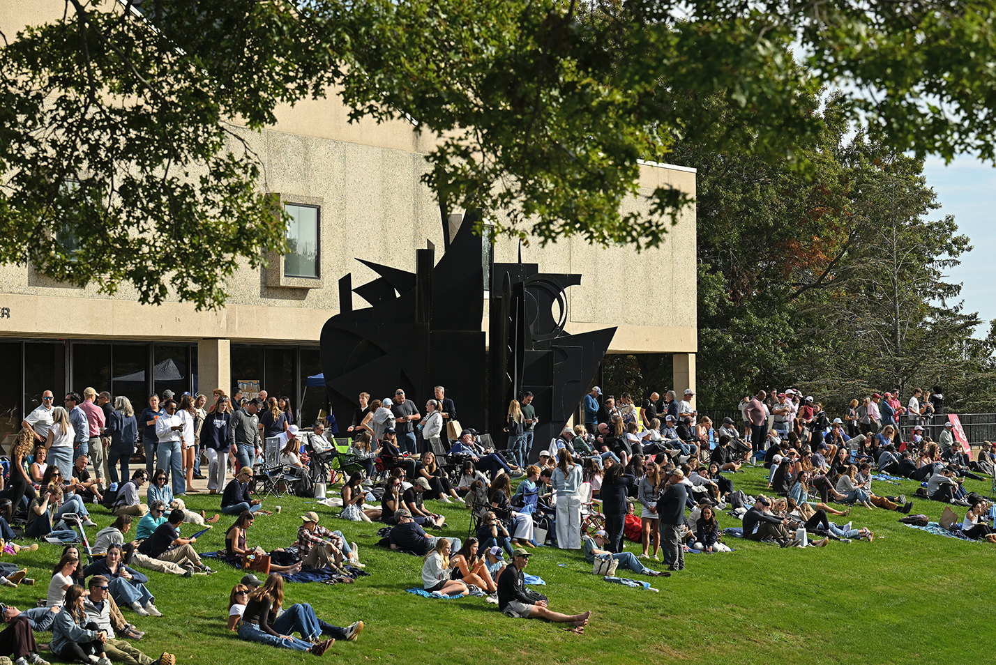 A crowd sits on a small bluff hillside in front of a brutalist concrete building with a large black steel sculpture.