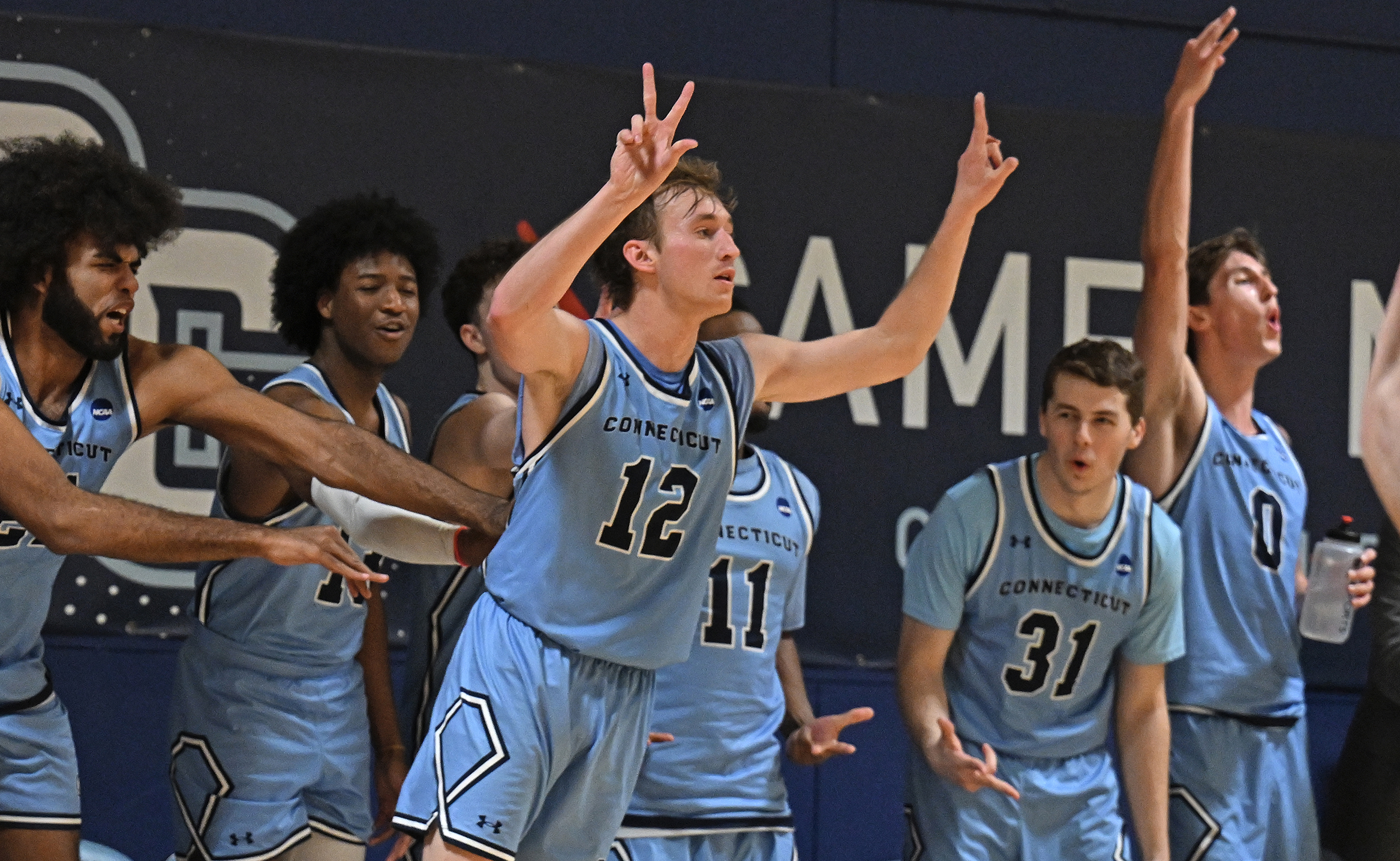 A basketball player raises his hands and three fingers to designate hitting a three-point field goal as his teammates on the bench behind him cheer.