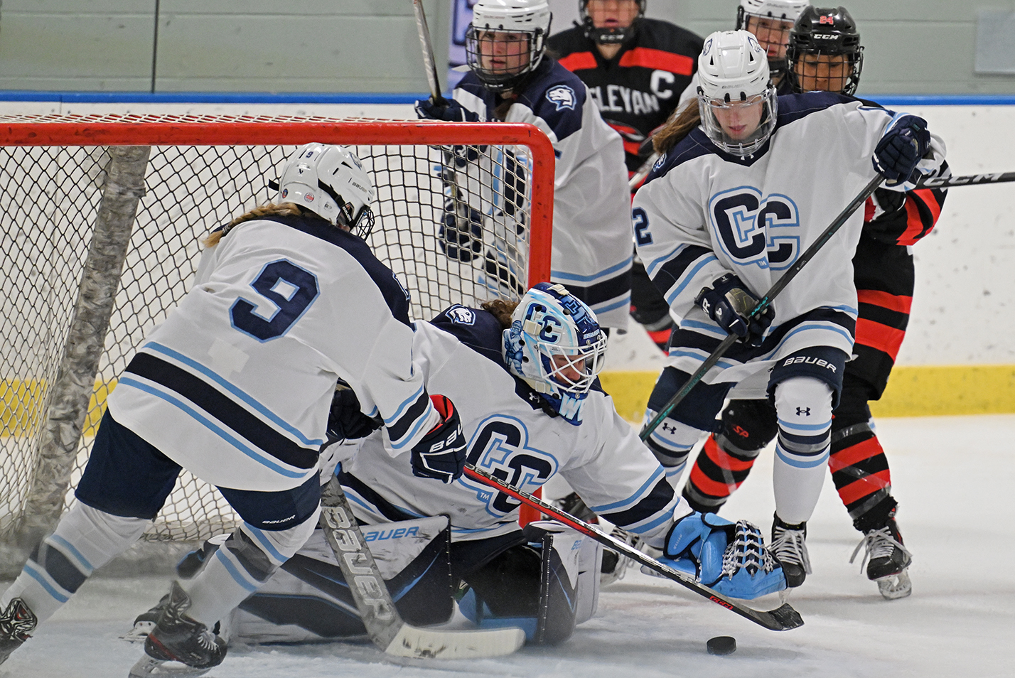 A college hockey goalie lunges forward to cover the puck as teammates and opponents swarm around the crease.