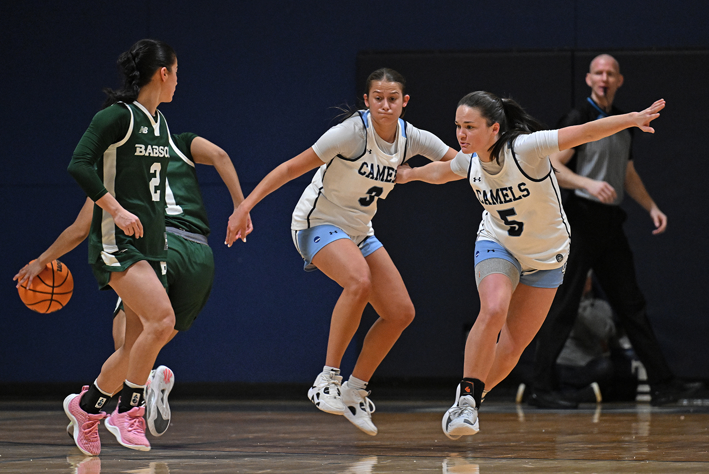 Two female basketball players in white uniforms and arms held wide set up on defense against players in green.