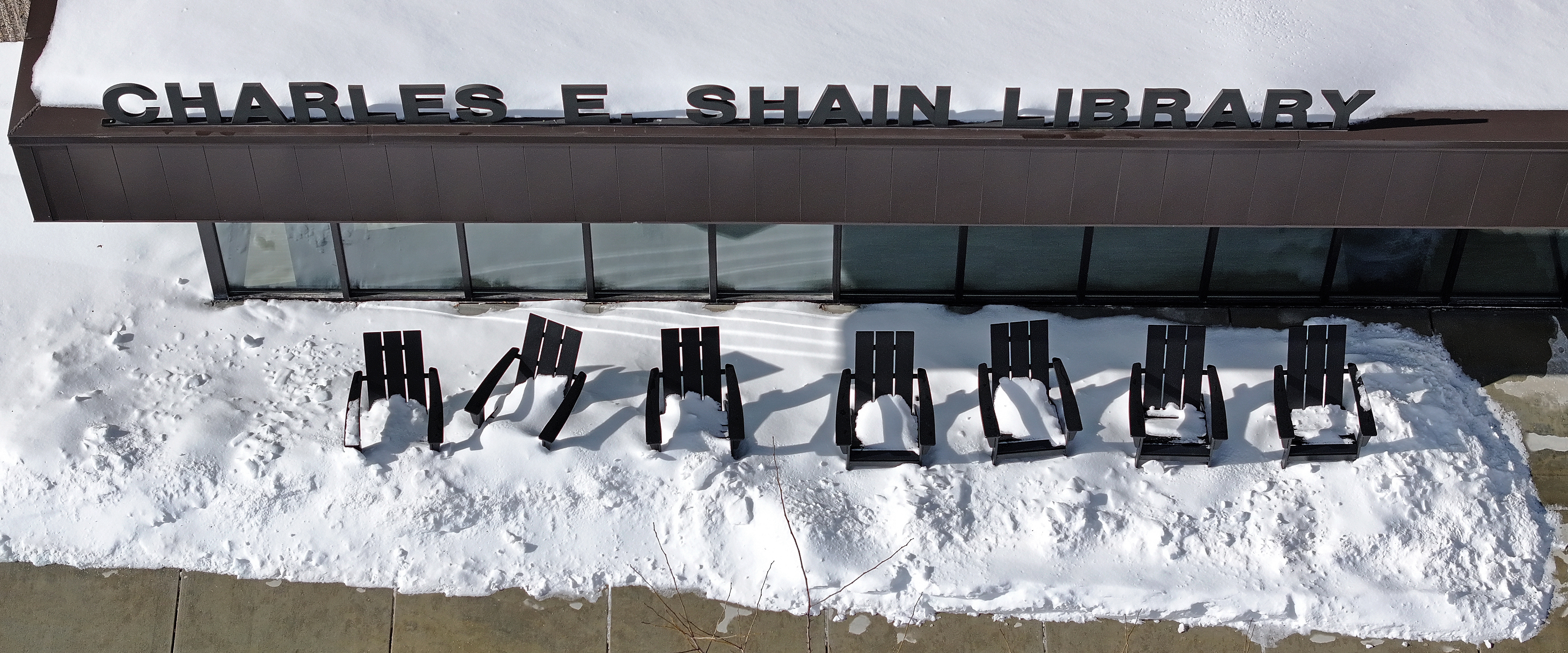 A row of Adirondack style chairs seen from overhead are partially buried in snow on a college library patio.