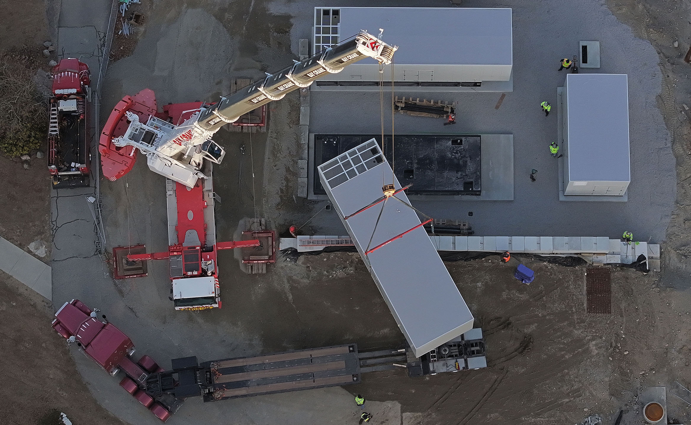 In a view from overhead, a large crane lifts a pre-fabricated building off a trailer and moves it to a nearby foundation.