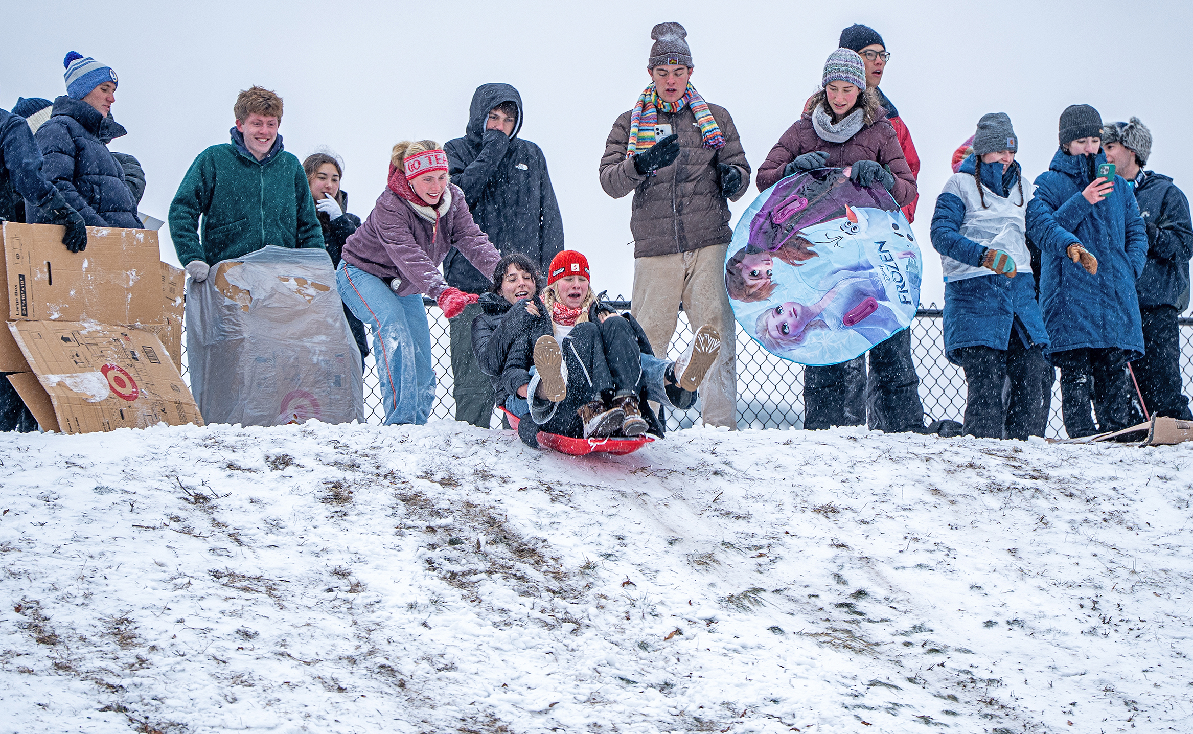 Two college students begin sledding down a snowy hillside as a crowd watches from the top of the hill.