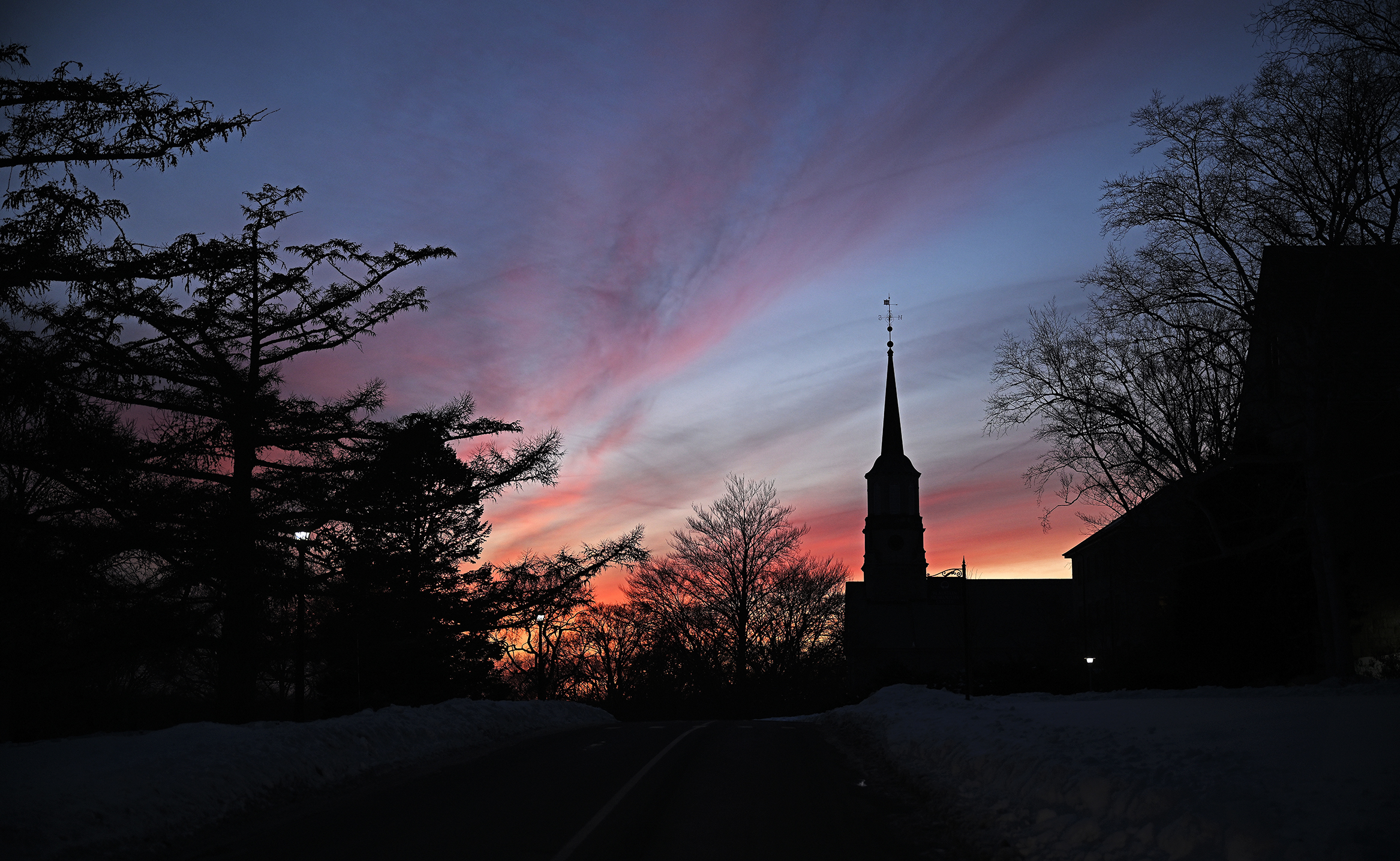College buildings, including a chapel steeple, are silhouetted against a brilliant red and purple sky at sunset.