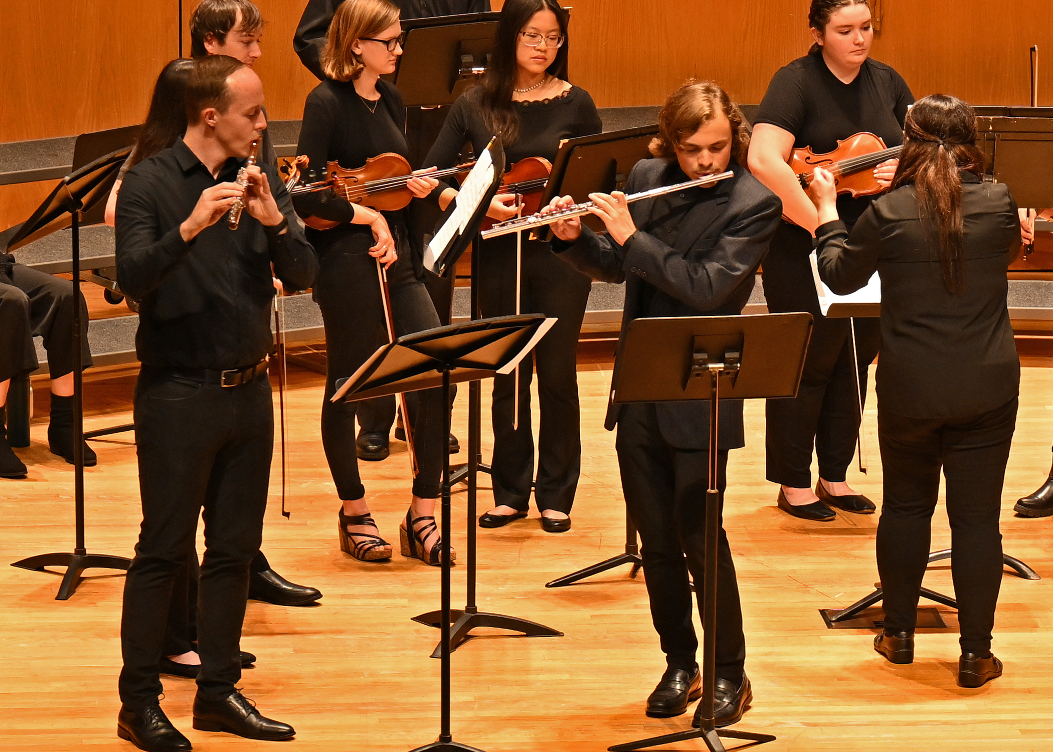 2 students each playing a flute, students with violins in the background