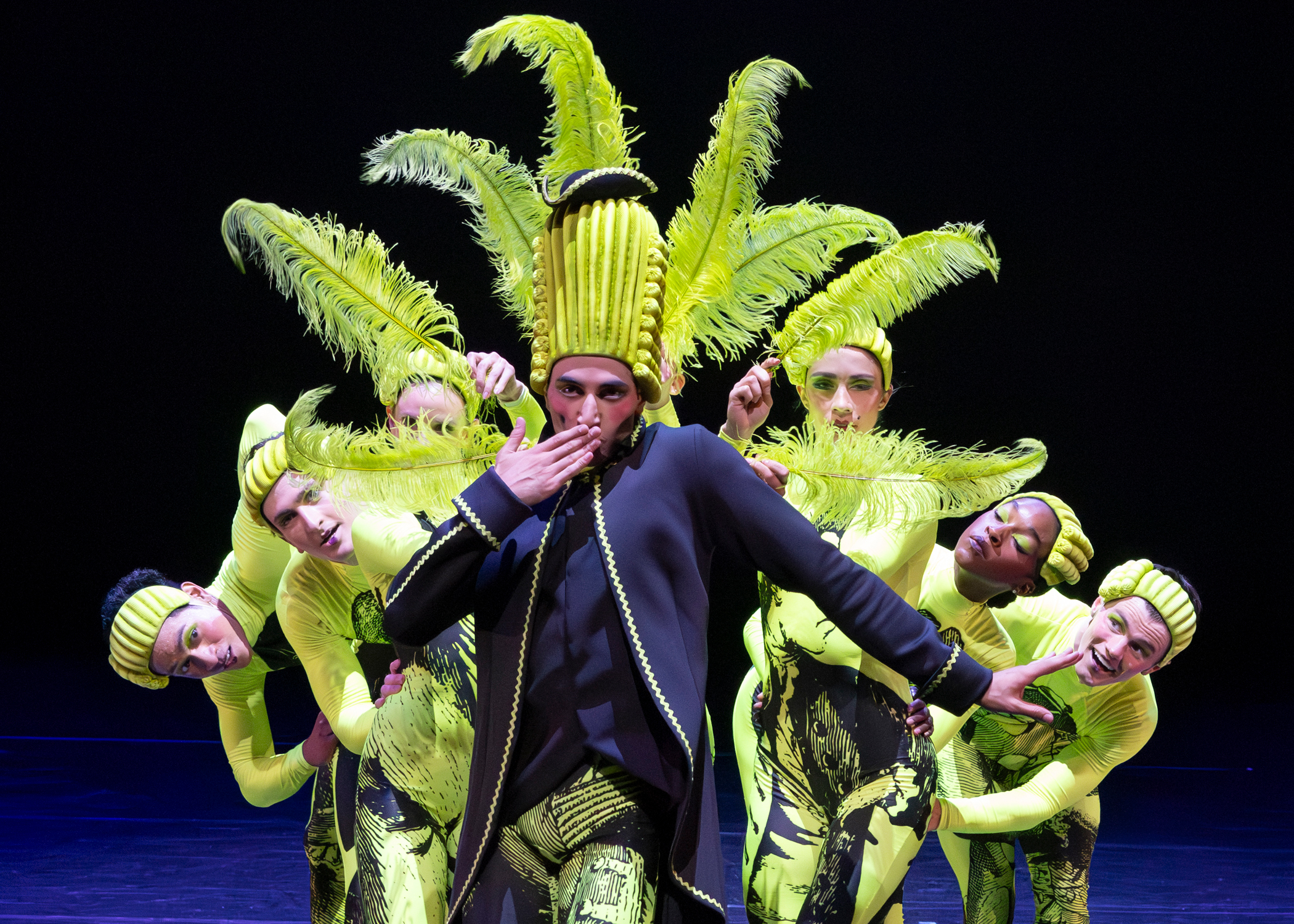 photo of 8 dancers, 7 wearing neon green bodysuits and holding green feathers surrounding a dancers in a black jacket and green 17th century wig
